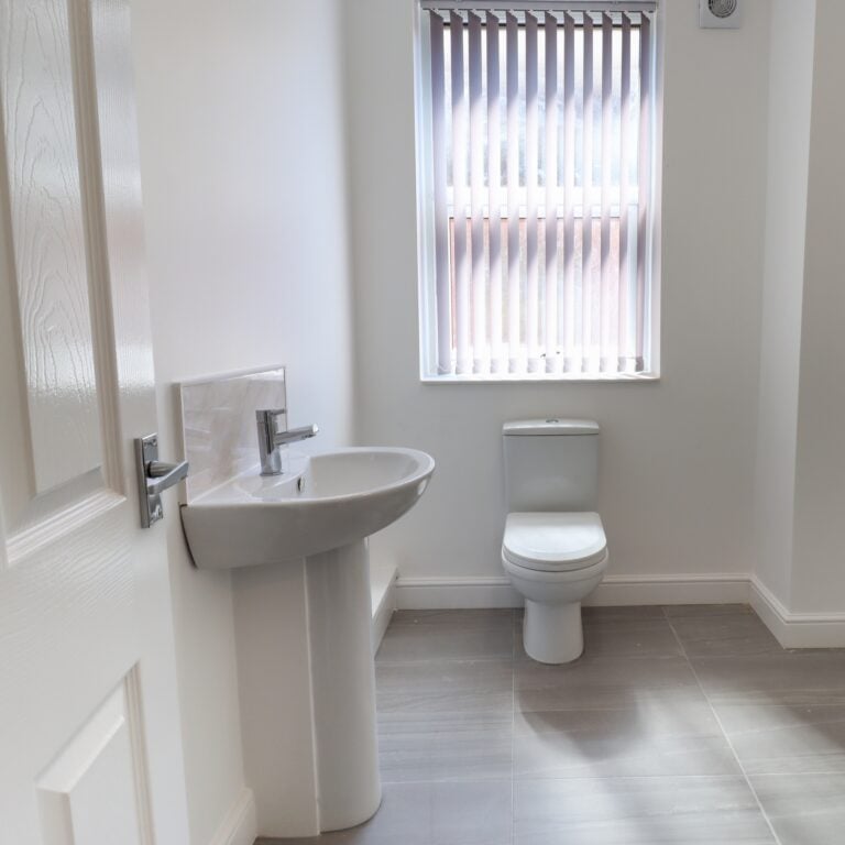 Photo of a modern brand new British bathroom showing a newly installed Toilet and Basin sink, with tiled grey floor, white walls and skirting boards with blinds up at the window