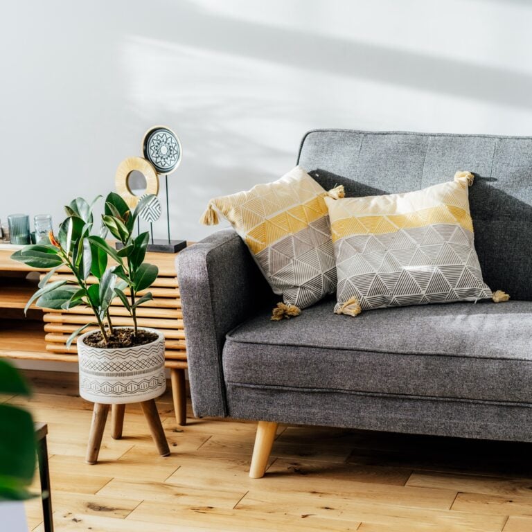 Stylish living room interior with designer console with decor and gray sofa with graphic pillows. White walls, green plants in the pots. Light wooden oak parquet. Modern Scandinavian style interior